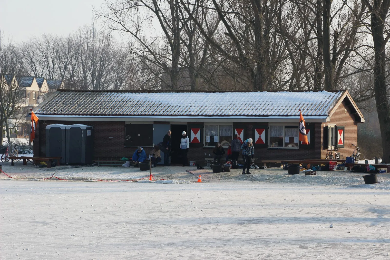 The brown clubhouse beside the frosted rink at Middelie, with bare trees behind, a portable toilet unit and people gathered at the door. — IJsclub De Ondersteuning