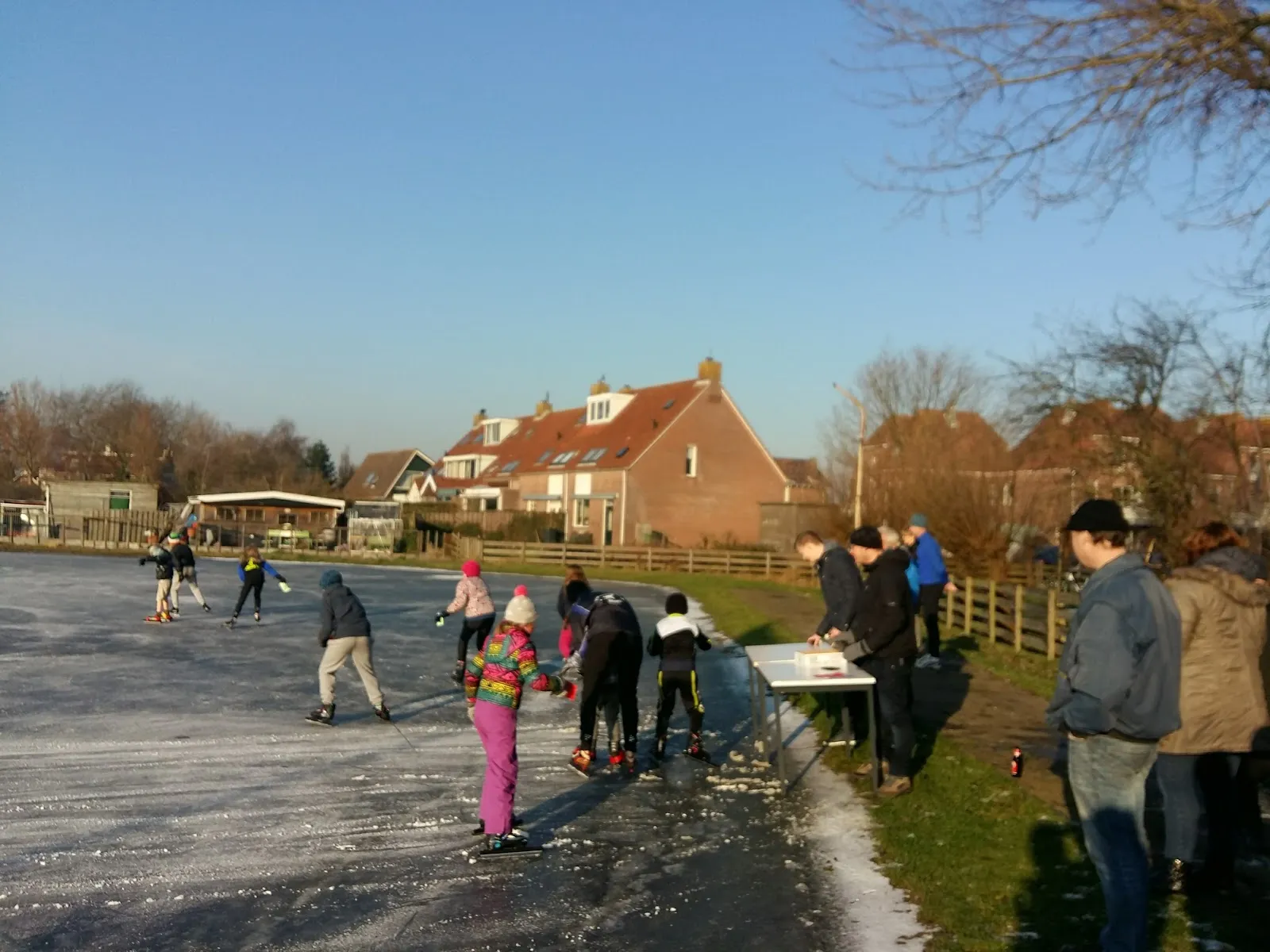 Skaters on the small frozen rink at Middelie on a sunny winter day, with a wooden fence and village houses with red roofs behind. — IJsclub De Ondersteuning