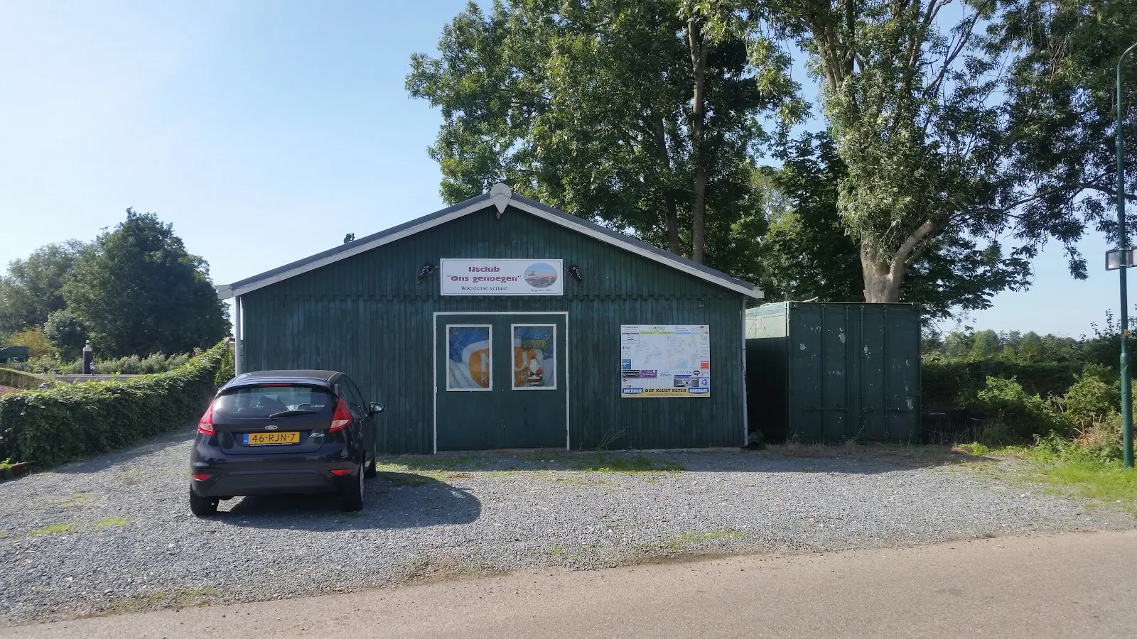 Het groene houten clubhuis van IJsclub de Toekomst in Bodegraven, met een auto op het grind ervoor en bomen erachter. – IJsclub de Toekomst