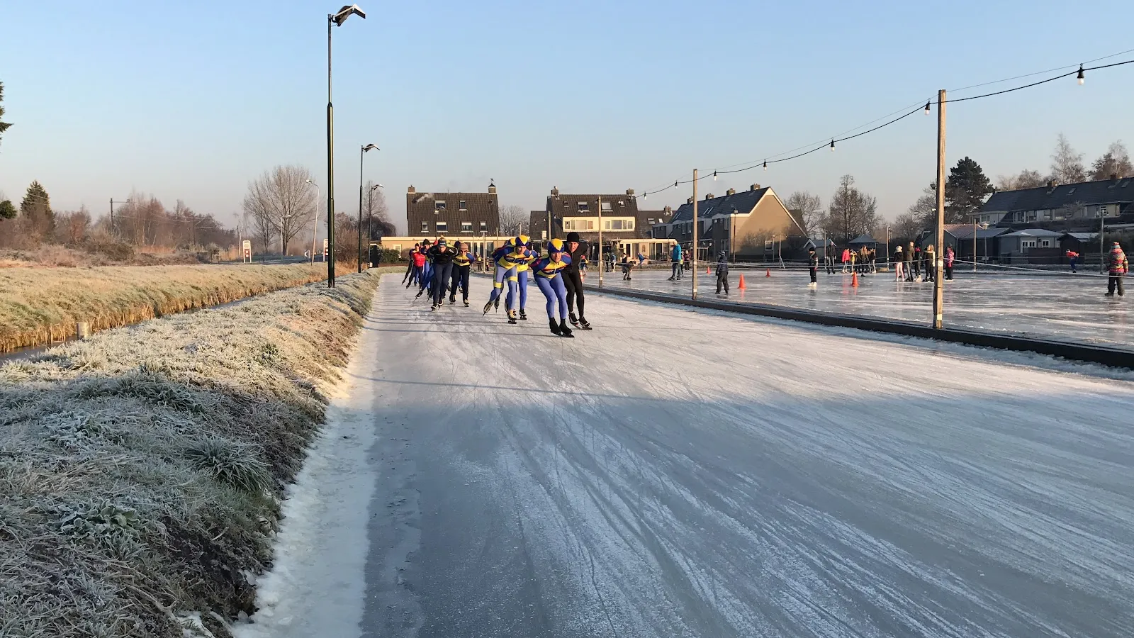 Schaatsers op de natuurijsbaan in Bodegraven op een vrieskoude ochtend, met lantaarnpalen en dorpshuizen achter de oever. – IJsclub de Toekomst
