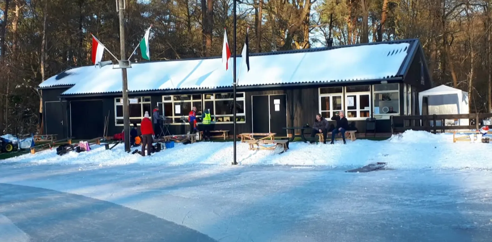 Donker houten clubgebouw met vlaggen in de sneeuw naast de baan, met een paar bezoekers buiten. – IJsclub Delden