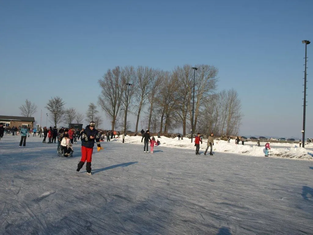 Schaatsers verspreid over de brede bevroren baan van Hellouw in de winterzon, met kale bomen en een lichtmast langs de overkant. – IJsclub D.E.S. Hellouw