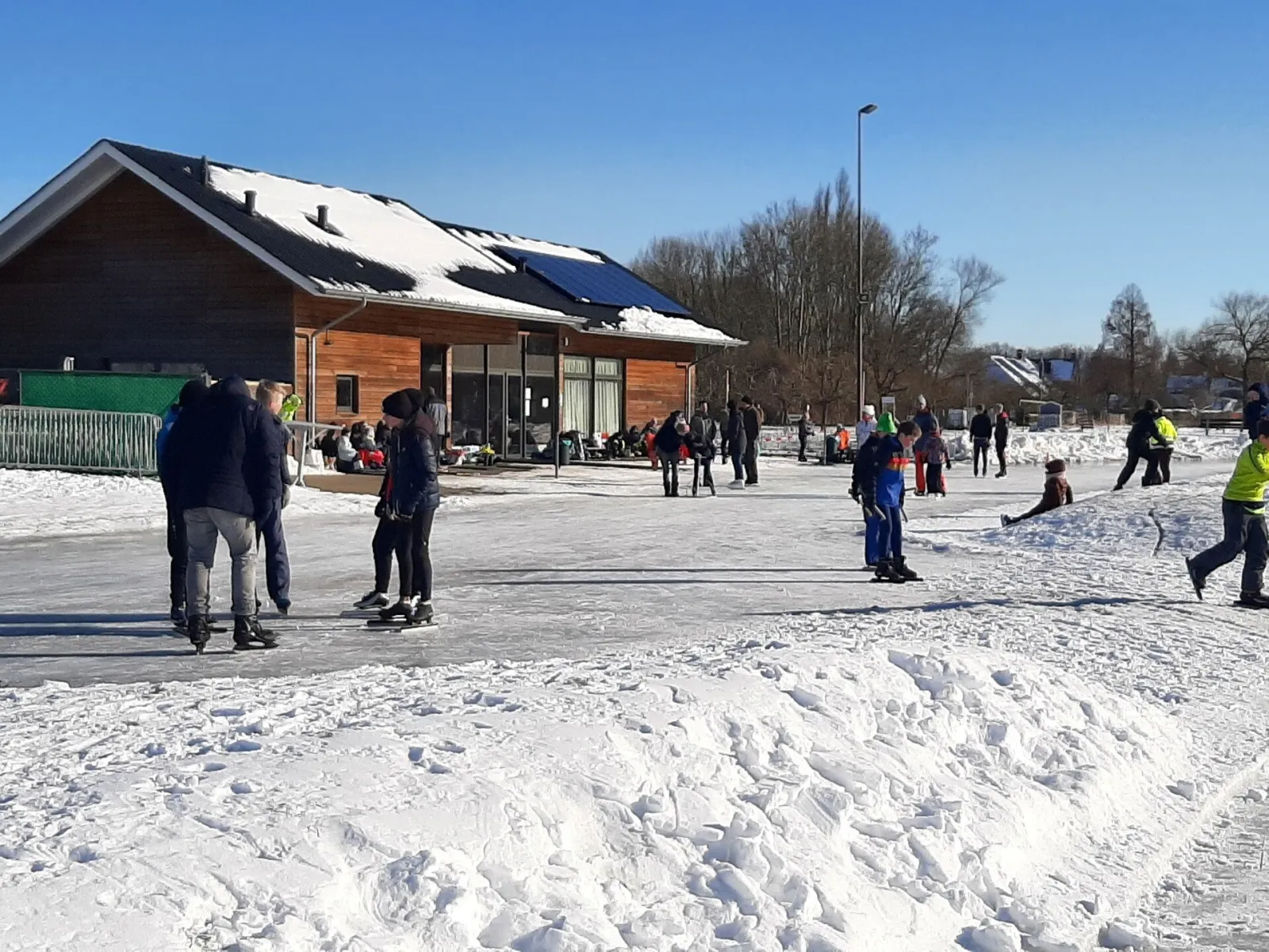 Het houten clubgebouw met zonnepanelen naast de besneeuwde baan van Herwijnen, met schaatsers en gezinnen op het ijs ervoor. – IJsclub d'n Ouwe Wiel