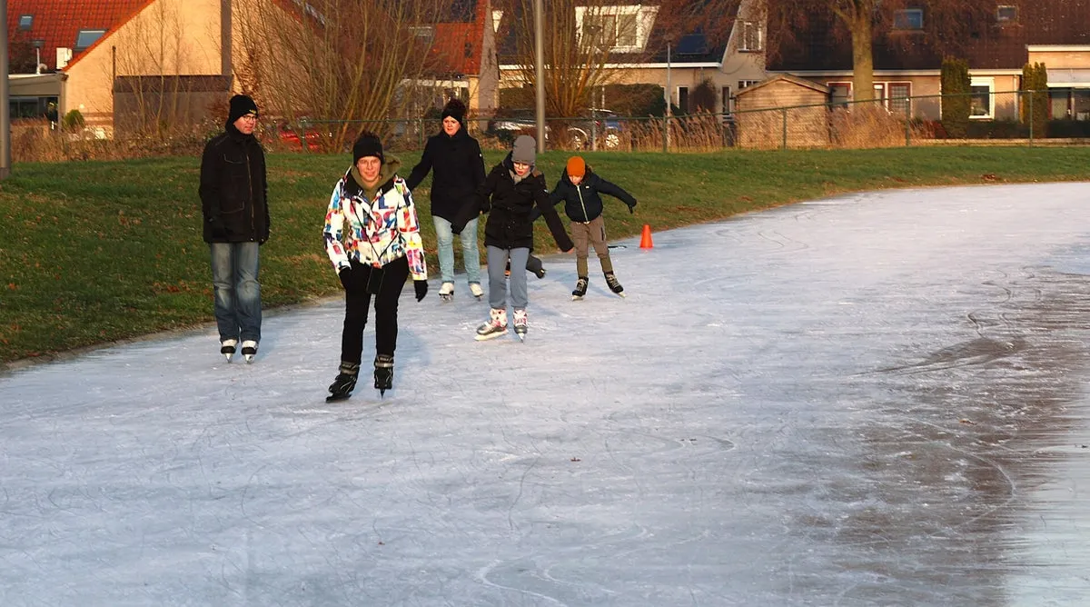 Schaatsers op de bevroren baan van Herwijnen op een heldere winterdag, met het dak van het clubgebouw, een afvalbak en huizen achter de baan. – IJsclub d'n Ouwe Wiel