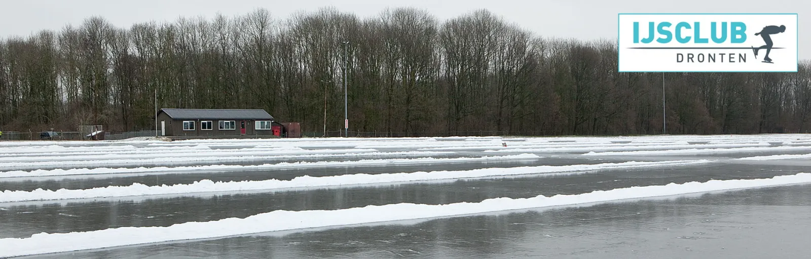 The low dark clubhouse of the Dronten natural-ice rink seen across ice that has been swept into lanes with banks of snow between them, with a row of bare trees behind. — IJsbaan Dronten (IJsclub Dronten)