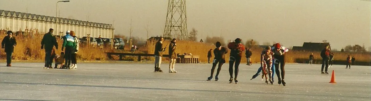 Skaters in small groups on the Wollebrand ice in low winter sun, with greenhouses and a pylon on the horizon. — IJsclub Eendracht (De Wollebrand)