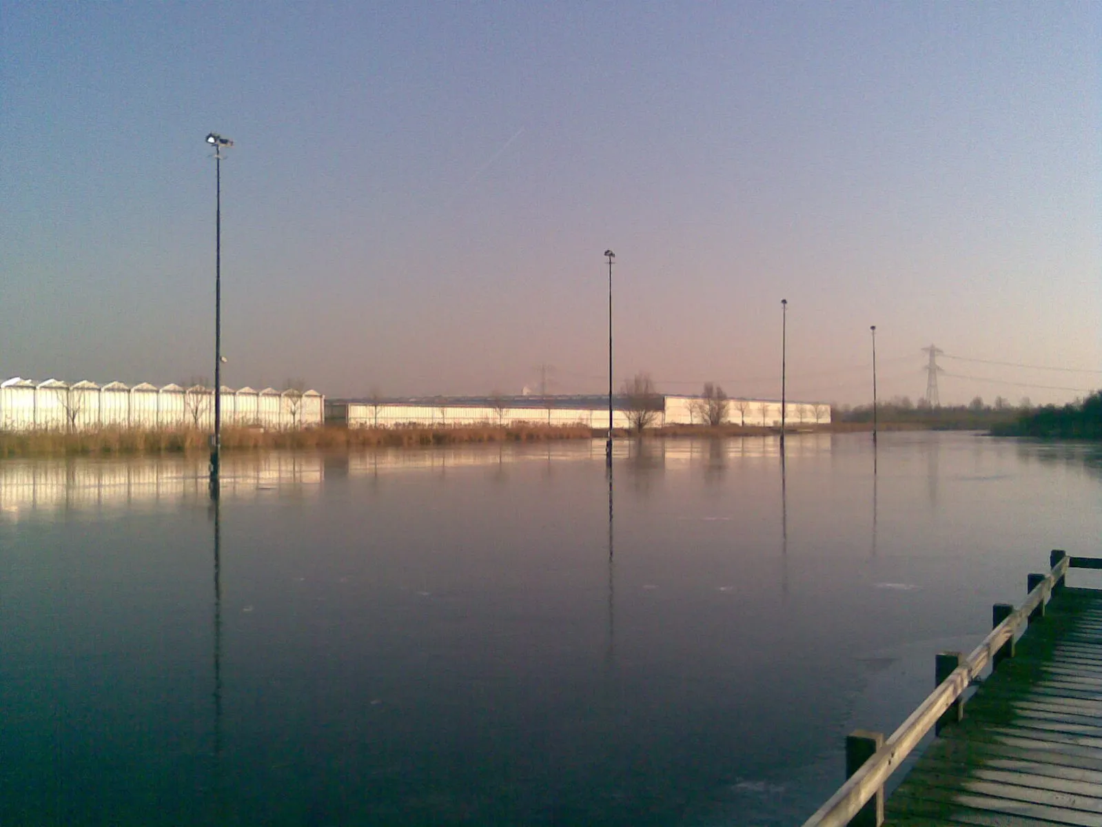 The frozen pond of the Wollebrand rink at dusk, with floodlight masts and greenhouses along the far bank and a wooden jetty in the corner. — IJsclub Eendracht (De Wollebrand)