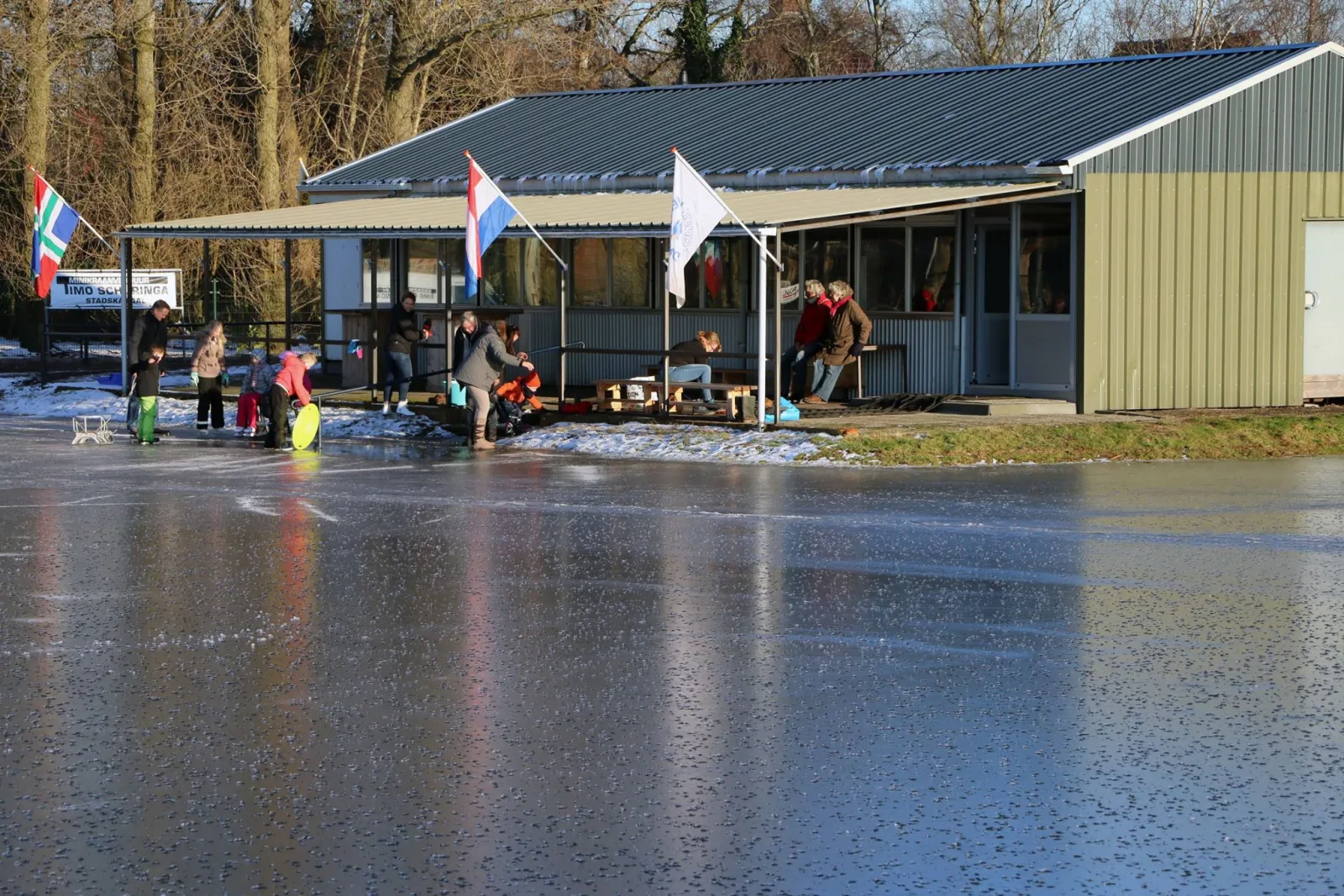 Het groene clubgebouw van IJsvereniging Eendracht met vlaggen en overdekt terras naast het ijs, met een paar schaatsers en bezoekers ervoor. – IJsbaan Eendracht (IJsvereniging Eendracht, Stadskanaal)