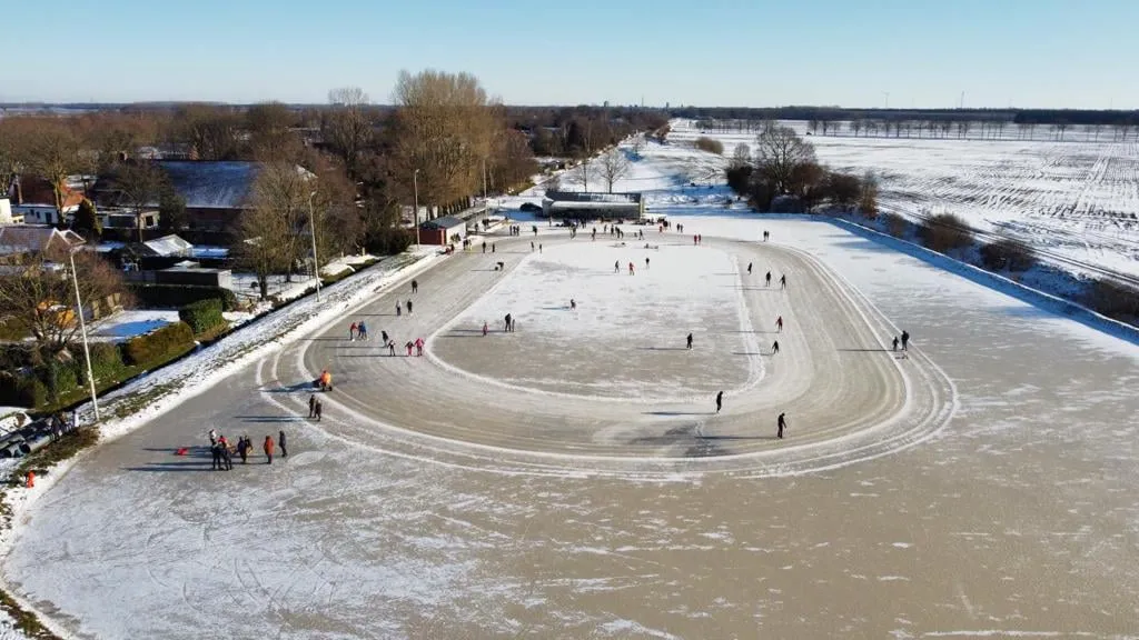 Luchtfoto van de ovale natuurijsbaan in Stadskanaal op een besneeuwde dag, met schaatsers verspreid langs de baan en het dorp en de velden eromheen. – IJsbaan Eendracht (IJsvereniging Eendracht, Stadskanaal)