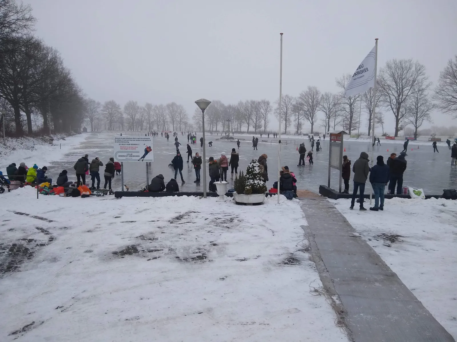 De baan van Eendracht in Vriezenveen onder de sneeuw, met mensen op het ijs, het toegangspad en vlaggenmasten aan de rand. – IJsclub Eendracht Vriezenveen