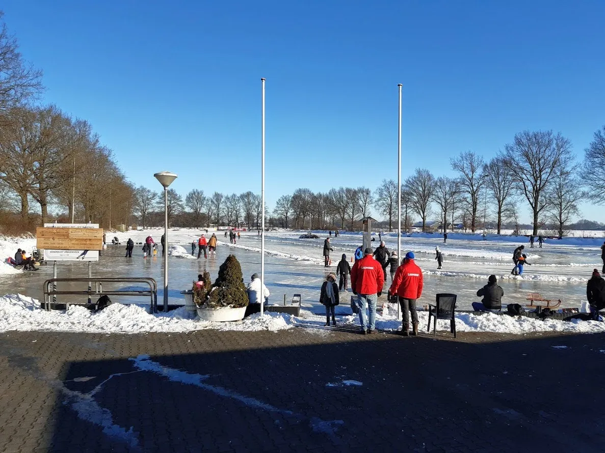 Schaatsers over het zonnige ijs in Vriezenveen met het toegangsplein op de voorgrond en twee vlaggenmasten naast de baan. – IJsclub Eendracht Vriezenveen