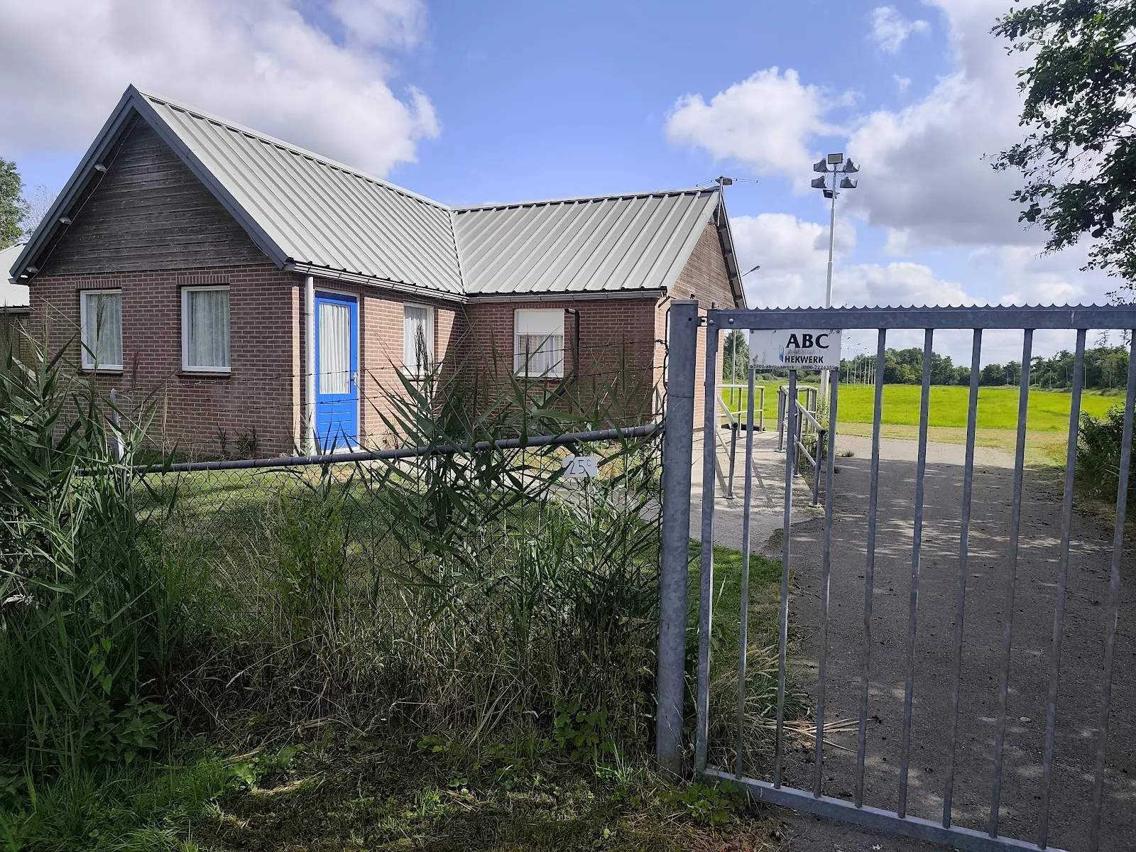 Bakstenen clubgebouw met een schuin metalen dak en een blauwe deur achter het metalen toegangshek van de baan. – IJsclub Excelsior