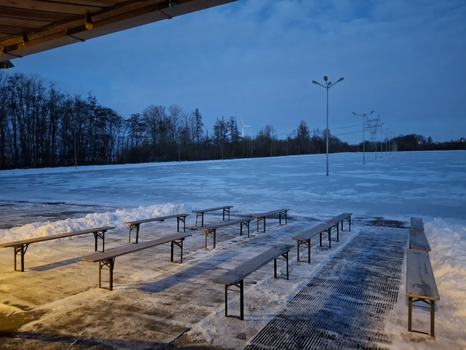 Lege banken onder het dak van het clubgebouw met uitzicht op de besneeuwde baan in de schemering, met lichtmasten en bomen op de achtergrond. – IJsclub Excelsior