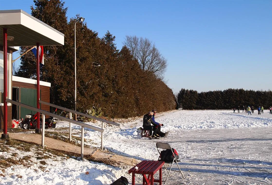 De veranda van het clubgebouw bij de baan in Gorredijk op een zonnige winterdag, met het besneeuwde ijs en een haag ernaast. – IJsclub Hâld Moed
