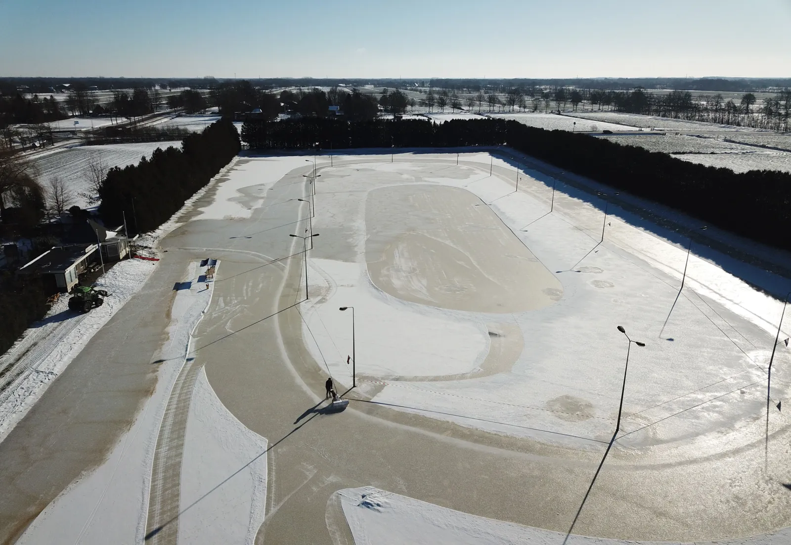 Luchtfoto van de bevroren, verlichte baan aan De Leijen in Gorredijk, met de baan afgetekend in het ijs en bomenrijen langs de randen. – IJsclub Hâld Moed