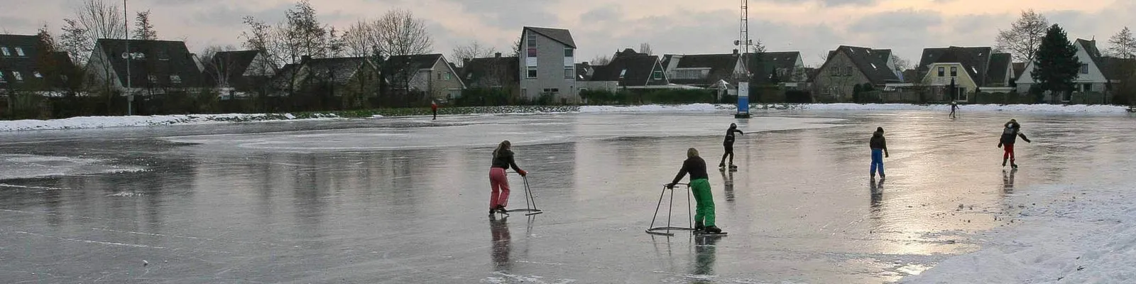 Schaatsers en kinderen met schaatsstoelen op het brede natuurijs van de landijsbaan in Hoogkarspel bij schemering, met langs de overkant de huizen van het dorp. – Landijsbaan Hoogkarspel (IJsclub Hoogkarspel)