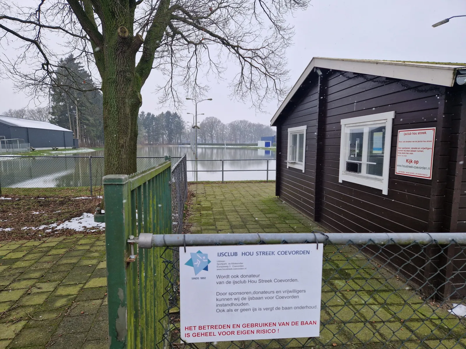The dark wooden clubhouse hut beside the flooded rink at Sportpark de Klinkenvlier, with the club's information sign on the fence and the blue sports hall in the distance. — IJsbaan Hou Streek (Coevorden)