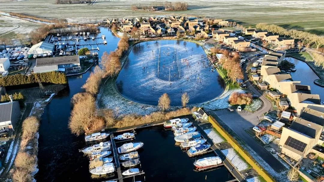 An aerial view of the oval ijsbaan in winter frost, surrounded by houses, a marina and the water channels around it. — IJsbaan Oldemarkt (IJsclub IJsvermaak Oldemarkt)