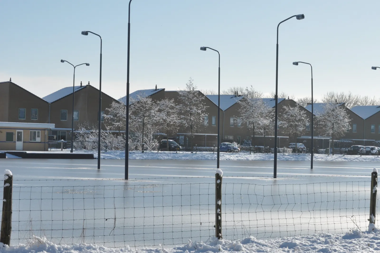 The frozen ijsbaan in winter behind a post-and-wire fence, with a row of tall lamp posts and houses along the far side. — IJsbaan Oldemarkt (IJsclub IJsvermaak Oldemarkt)