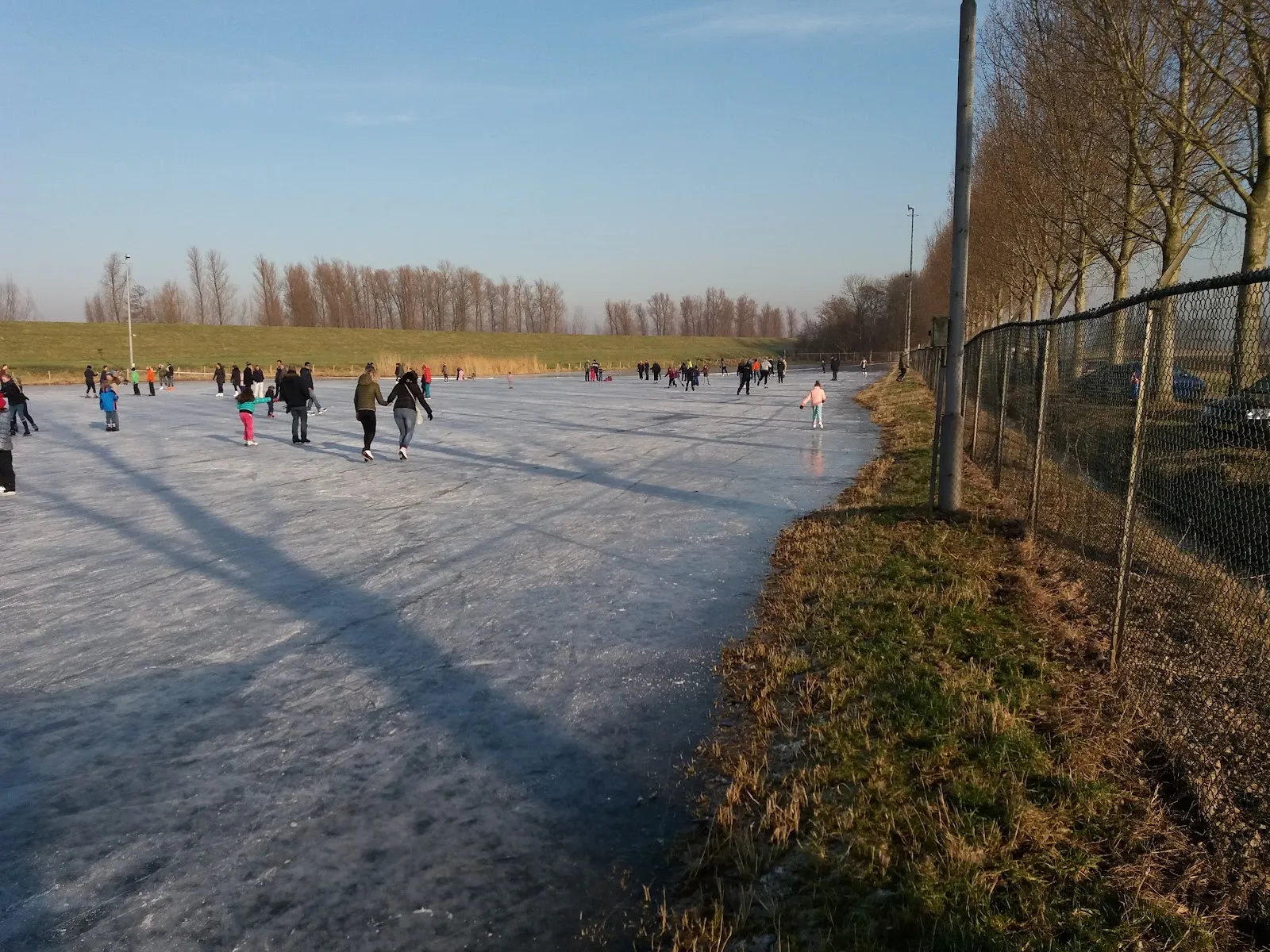 A long stretch of the Lage Zwaluwe natural-ice rink with a few skaters far off and a fence along the bank. — IJsbaan Lage Zwaluwe (Koninklijke IJsclub)