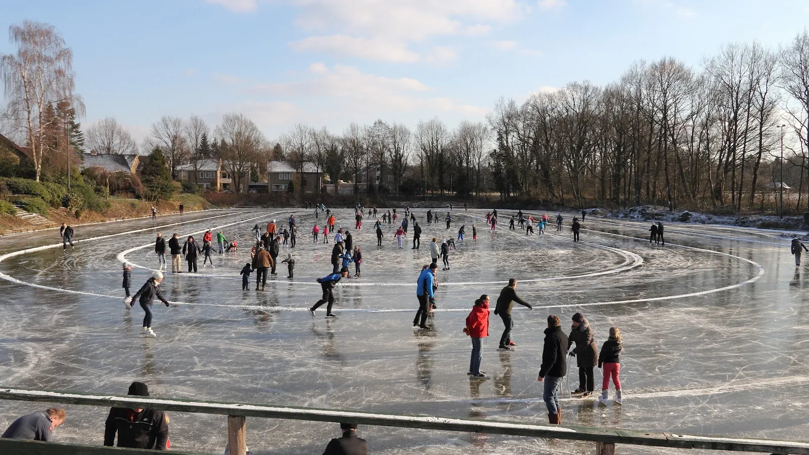 Schaatsers verspreid over de bevroren vijver van Lonneker met de ovaal afgetekend in het ijs, huizen en kale bomen langs de overkant. – IJsclub Lonneker
