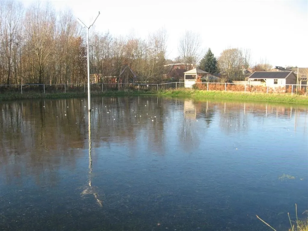 The flooded rink field at Marum West in winter light, a single lamp post standing in the shallow water with houses and bare trees along the far edge. — IJsclub Marum West