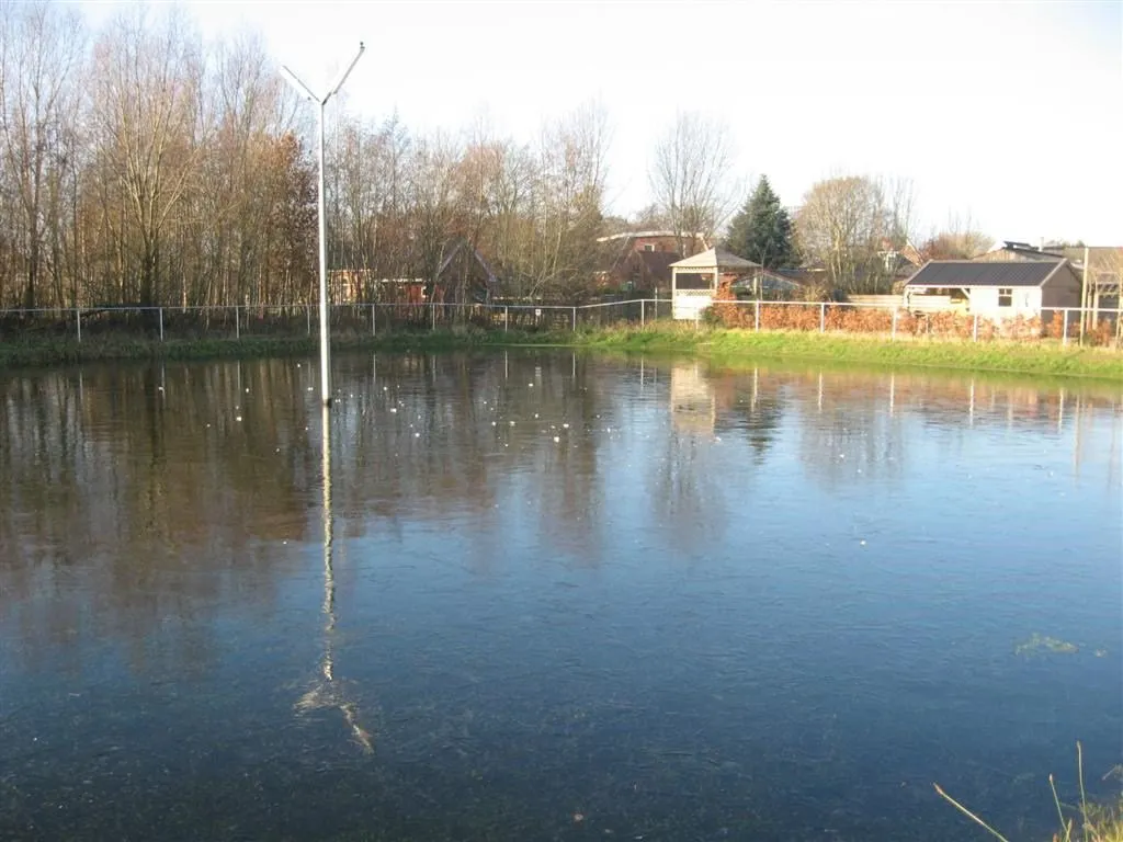 The frozen Marum West rink field with a floodlight mast, trees and a clubhouse roof beyond the fence. — IJsclub Marum West