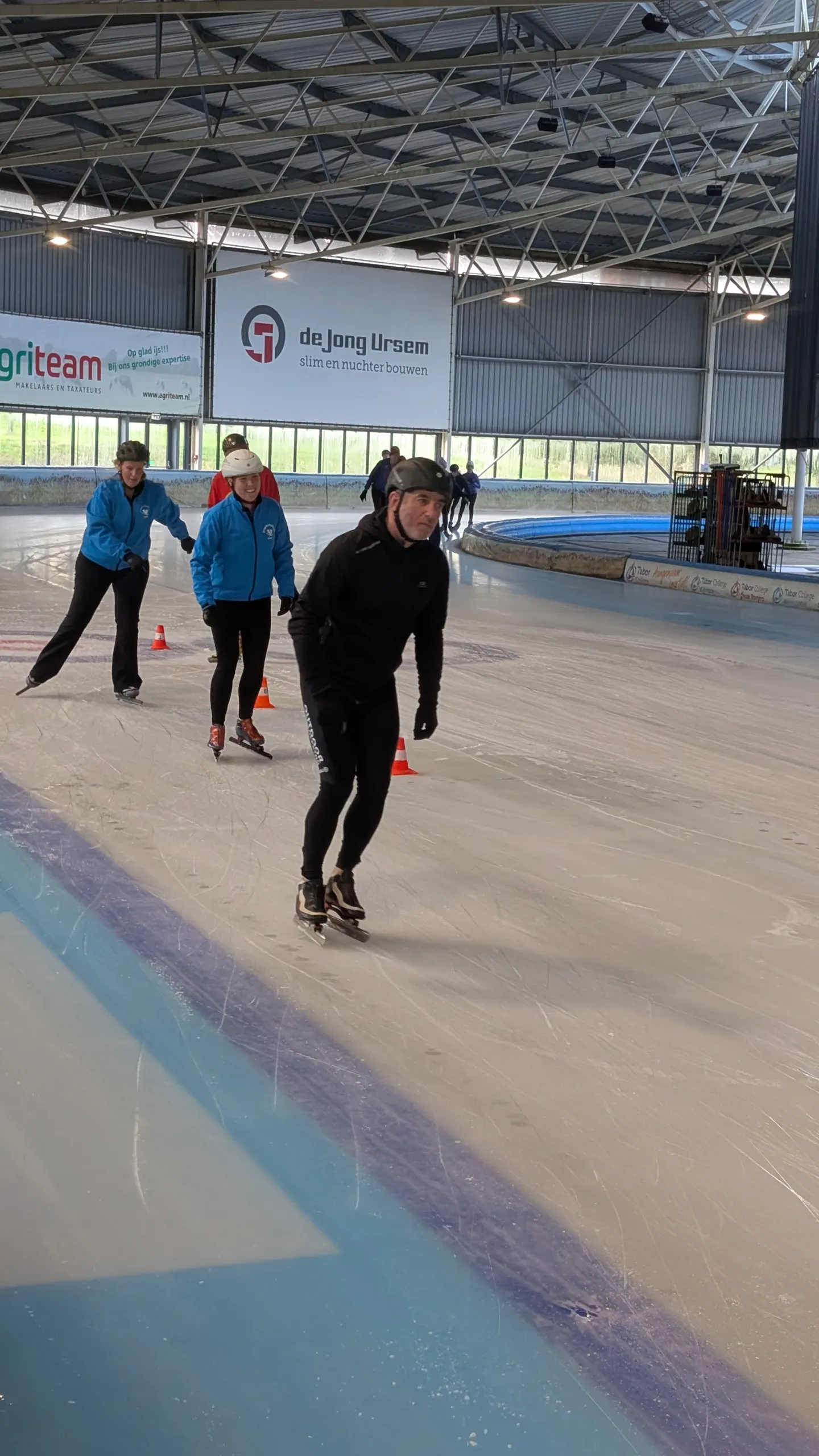 Skaters on the covered ice of the Nibbixwoud ice club's rink, with the roof and the sponsor boards behind them — IJsbaan Nibbixwoud (IJsclub Nibbixwoud)