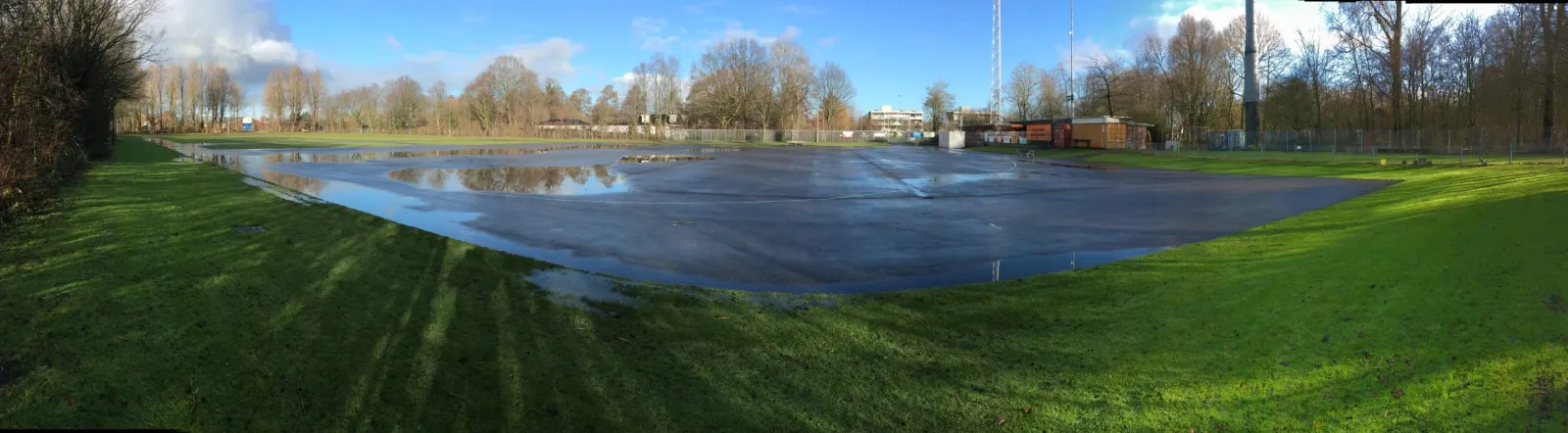 Een panorama van het terrein van de baan in Nieuw-Vennep in de winter van 2021, het onder water gezette ijsvlak omzoomd door gazon, bomen en de paal van de toegangspoort. – IJsclub Nieuw-Vennep en Omstreken