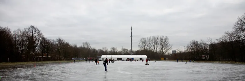 Een breed beeld van schaatsers op het grijze natuurijs van de baan in Nieuw-Vennep, met een witte tent en kale bomen aan de overkant onder een bewolkte lucht. – IJsclub Nieuw-Vennep en Omstreken