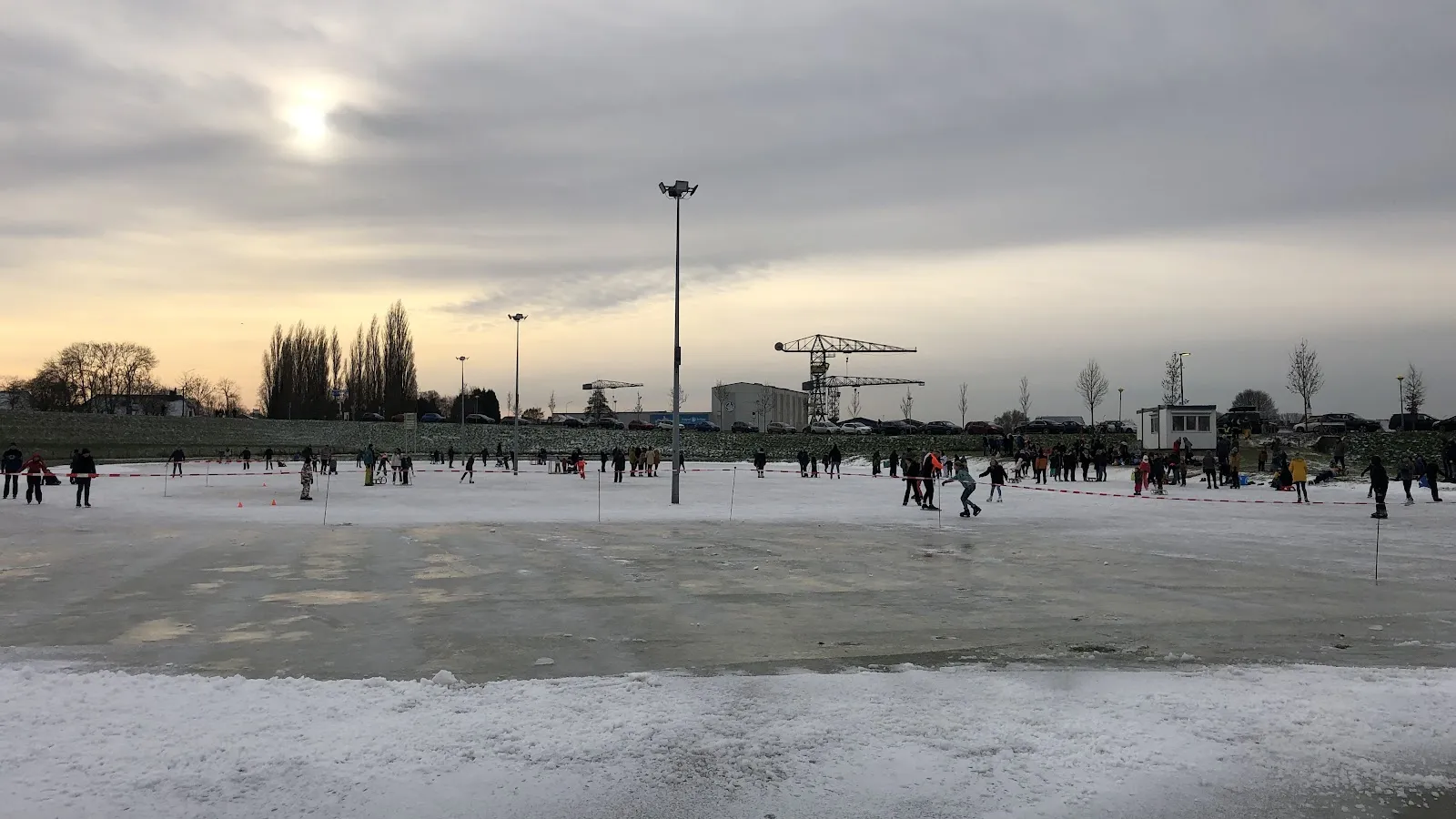 Een lage winterzon boven de brede baan van Gendt, met schaatsers langs de verre rand, een lichtmast en een kraan erachter. – IJsclub Nooitgedacht Gendt
