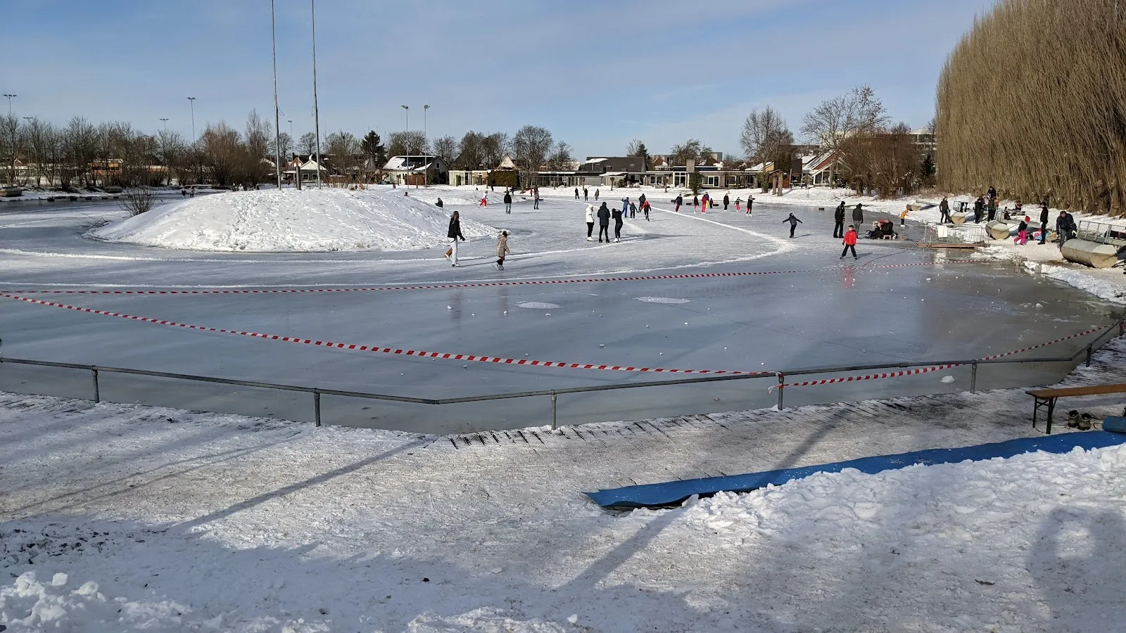 Schaatsers op de natuurijsbaan van Gendt op een besneeuwde zonnige dag, met een sneeuwberg op het ijs, een omrande rand en dorpshuizen erachter. – IJsclub Nooitgedacht Gendt