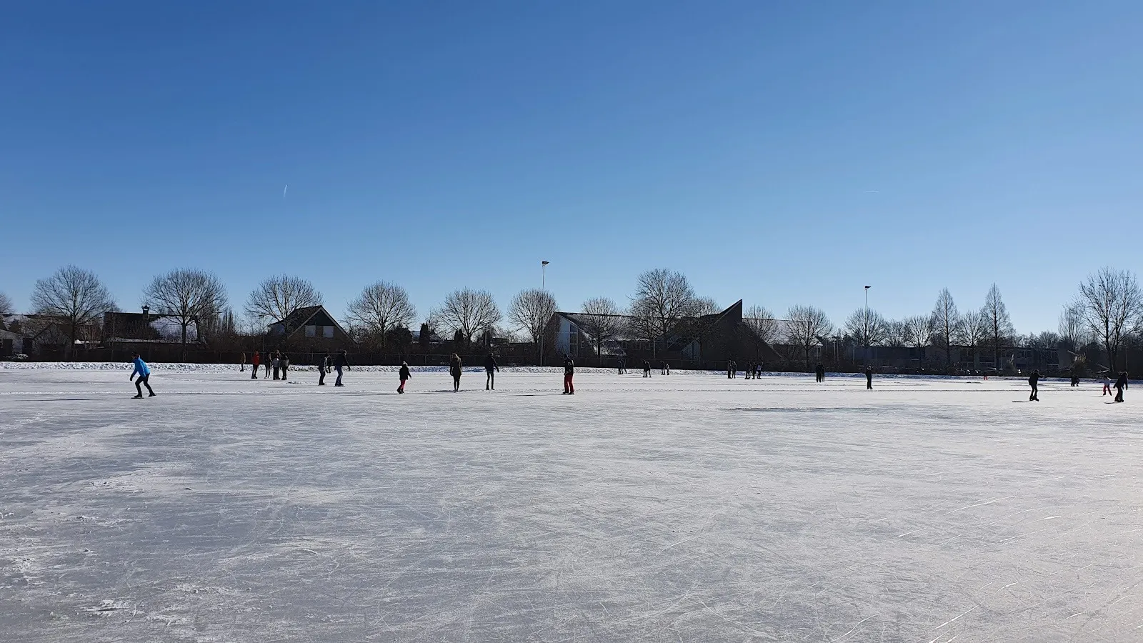 Schaatsers verspreid over het brede, heldere ijs van de baan in Oudenhoorn onder een strakblauwe lucht, met boerderijen en kale bomen langs de overkant. – IJsclub Nooitgedacht Oudenhoorn