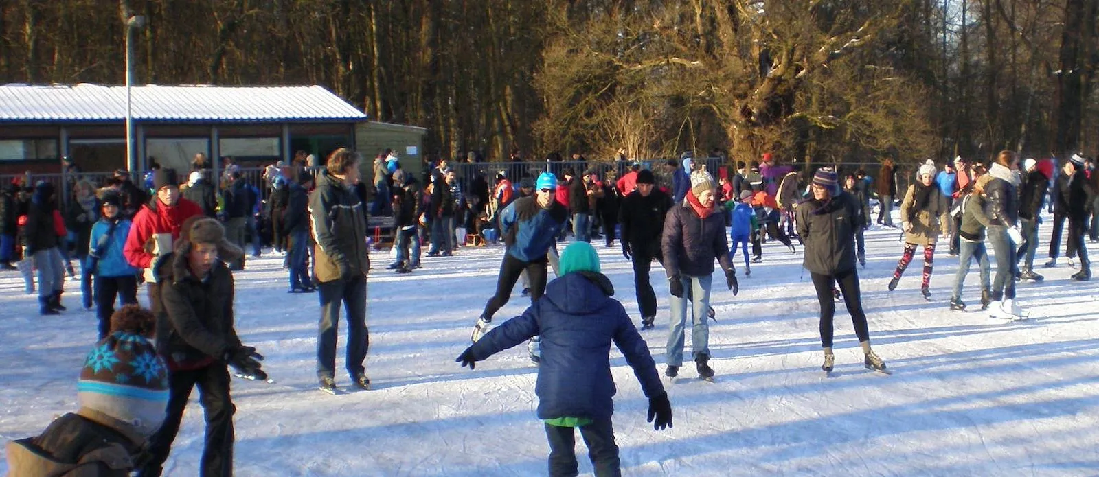 Crowd of skaters on the sunlit ice at the Oegstgeest rink, with the clubhouse and old trees behind. — IJsclub Oegstgeest