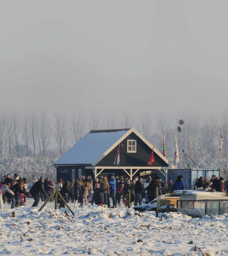 Het donkere houten clubgebouw van de baan in Werkendam op een vrieskoude dag, met de Nederlandse vlag, clubvlaggen en een luidsprekermast, en een menigte schaatsers langs het hek ervoor. – IJsbaan Werkendam (IJsclub Olympia)