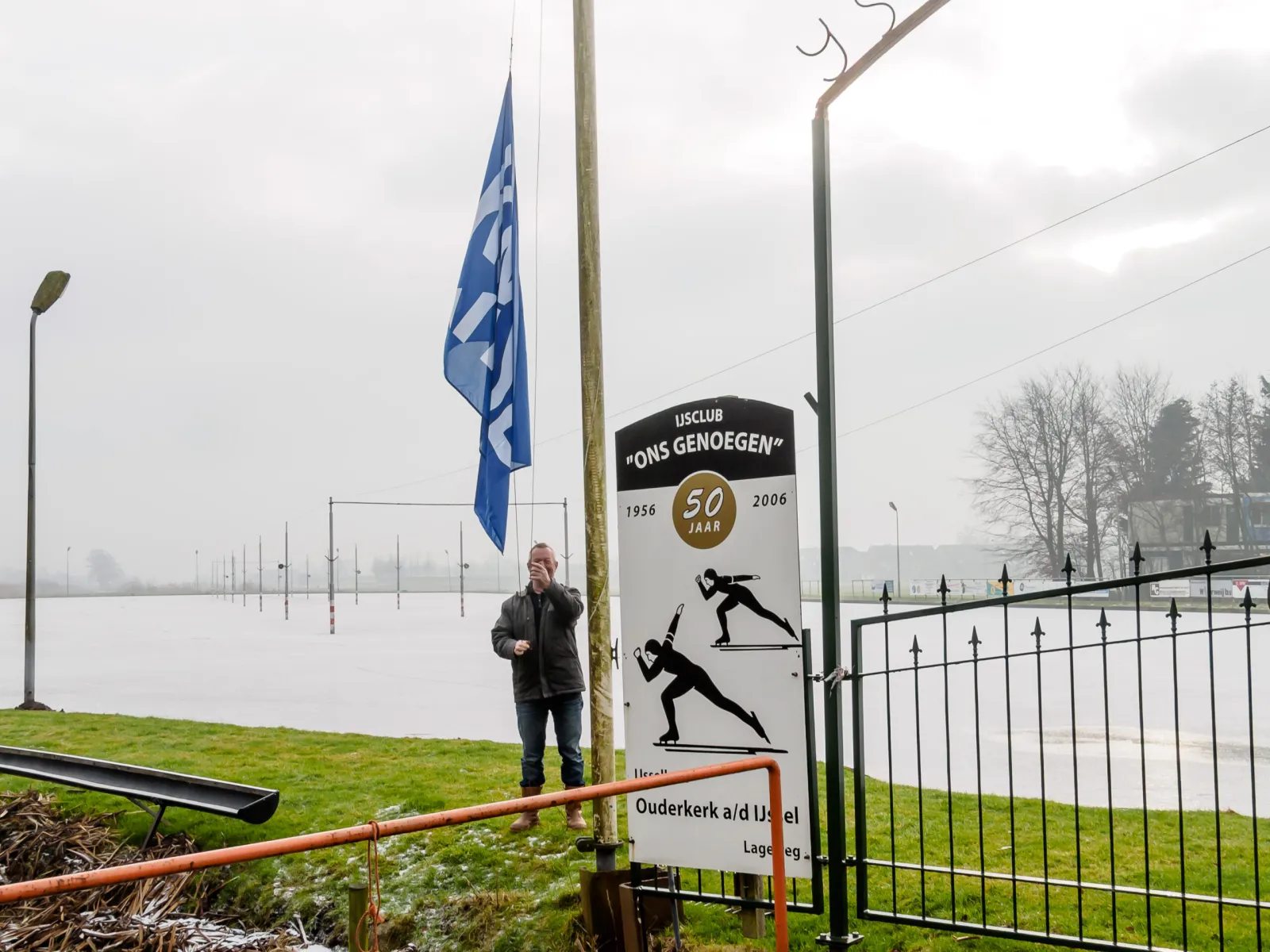 The rink's entrance sign for IJsclub Ons Genoegen (1956-2006, 50 years) beside the iced field, with a flag being raised and a row of lighting poles in the mist. – IJsbaan Ons Genoegen (Ouderkerk aan den IJssel)