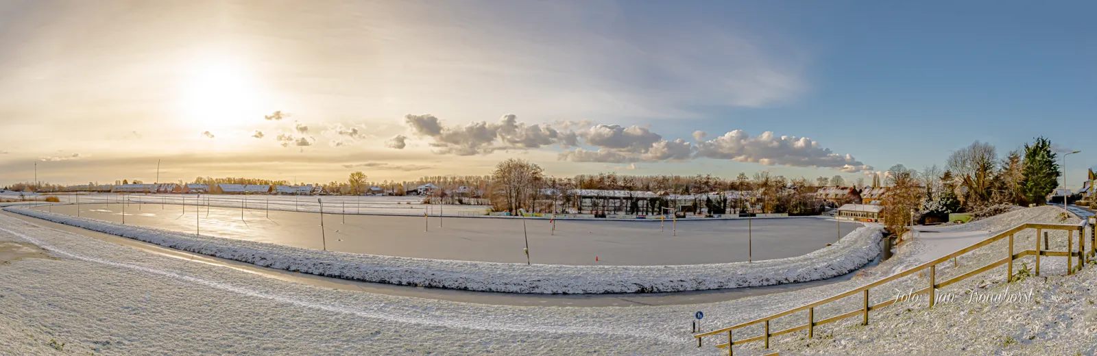 Wide winter panorama of the frozen field rink at the IJssellaan, with its floodlight poles, the snow-covered bank in front and the clubhouse and houses along the far side under a low sun. – IJsbaan Ons Genoegen (Ouderkerk aan den IJssel)