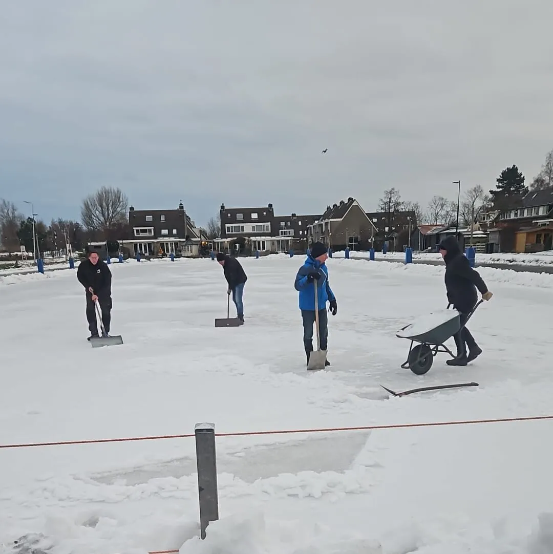 Volunteers clearing snow from the natural ice on the Boskoop combi track in January 2026, with shovels and a wheelbarrow on the white surface and village houses along the edge. — Combibaan IJsclub Otweg (Boskoop)