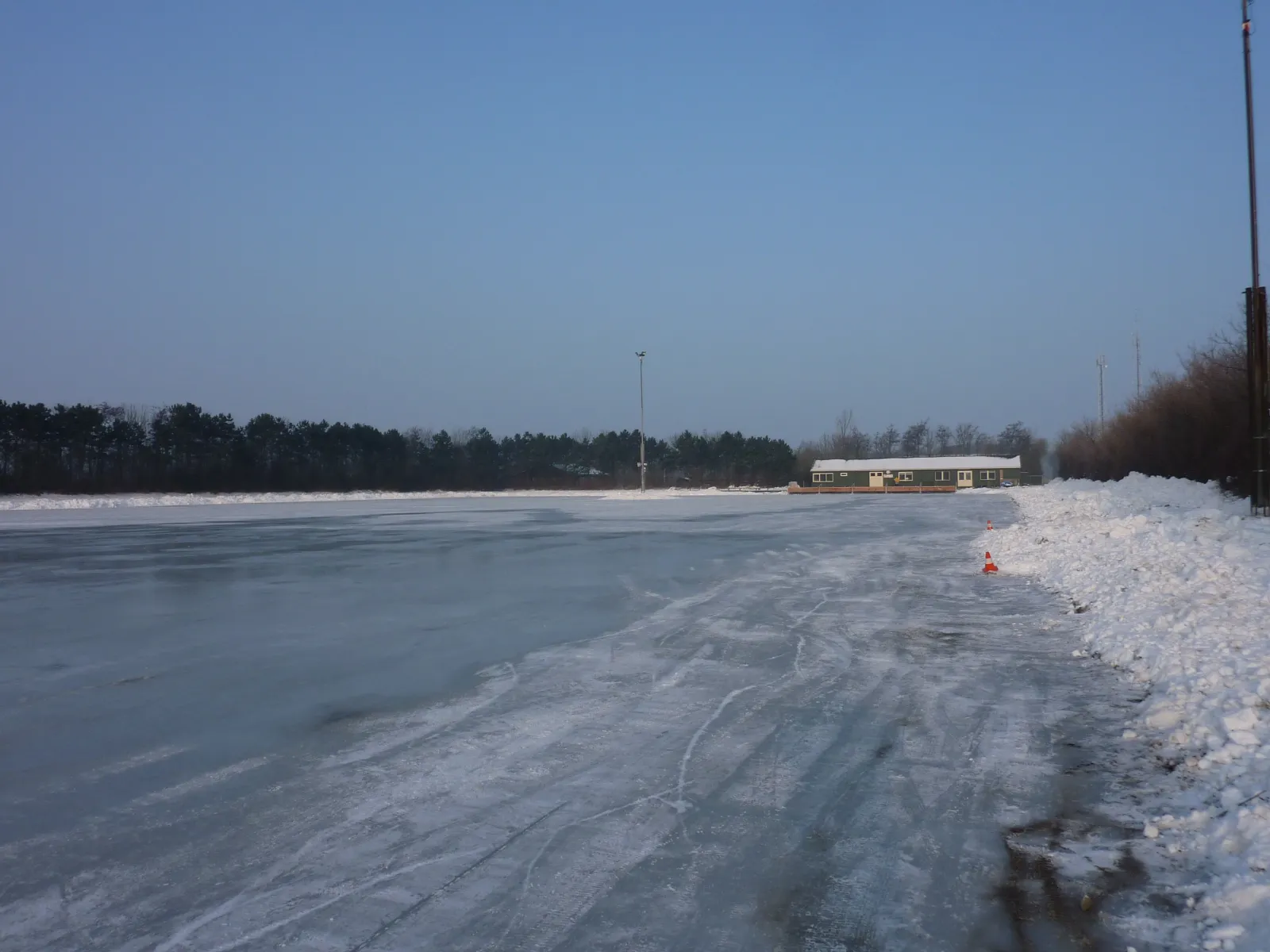 De lege natuurijsbaan van Ouddorp onder een heldere winterlucht, met sneeuwwallen langs de rand, een lichtmast en het groene clubgebouw erachter. – IJsclub Ouddorp