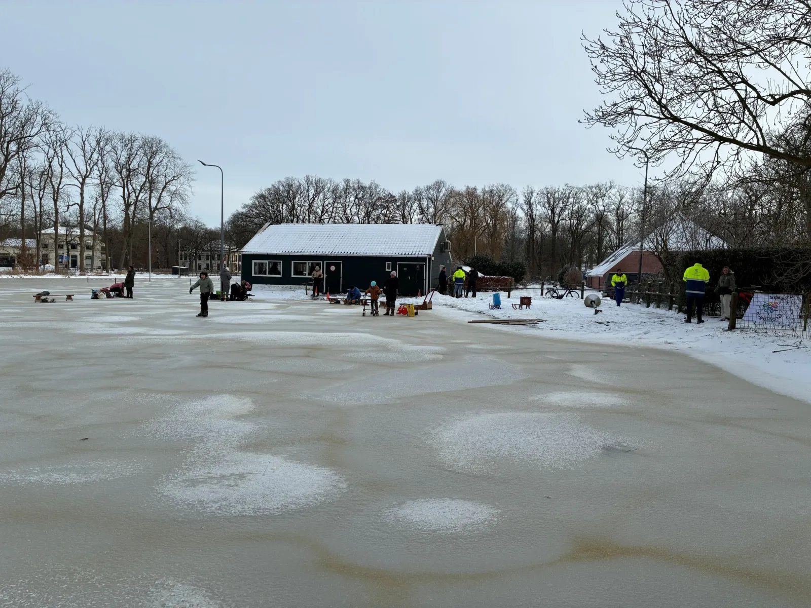 Donker houten clubgebouw met een besneeuwd dak aan de rand van het ijs, met een paar vrijwilligers in de verte. – IJsclub Slochterbosch