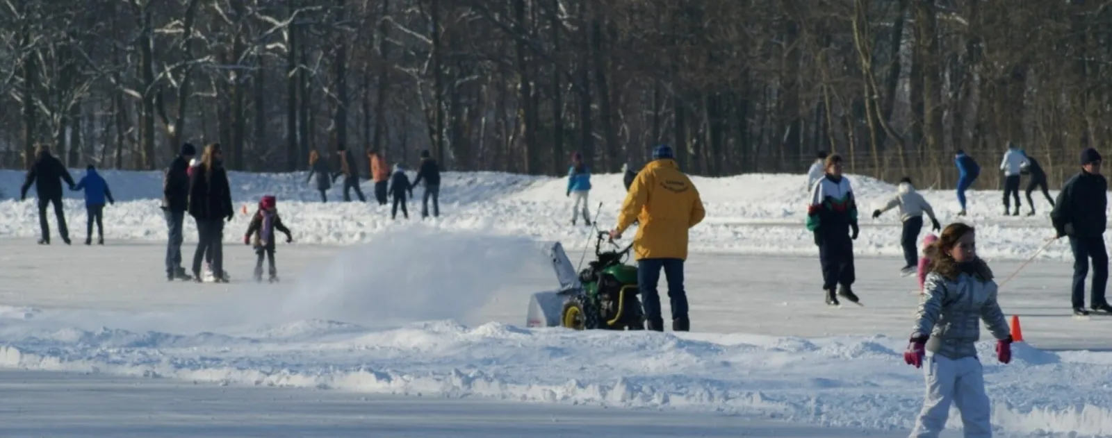Skaters on the swept natural-ice rink in Swifterbant on a bright winter day, with a driver clearing the ice with a small sweeper and bare woods behind. — IJsbaan Swifterbant (IJsclub Swifterbant)