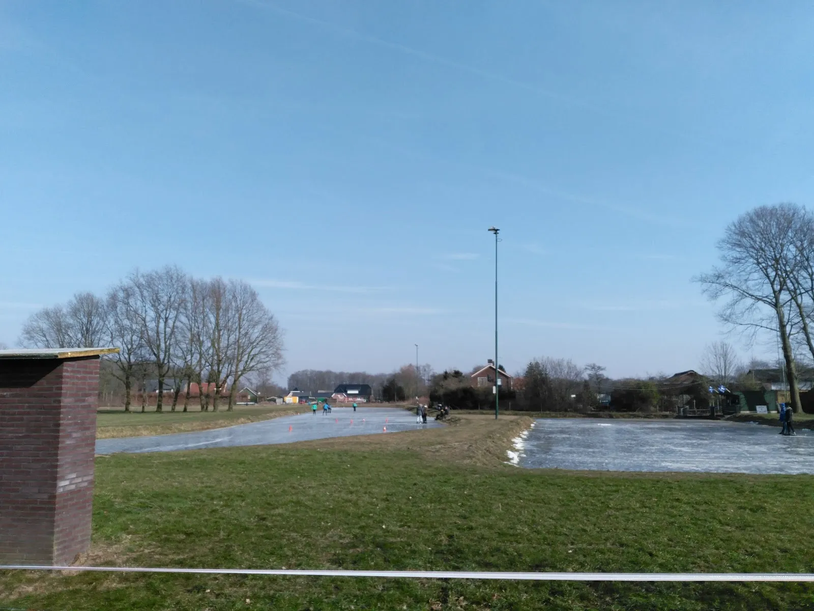 Schaatsers in de verte op de twee armen van de bevroren baan van Boesveld onder een wijde blauwe lucht, met een lichtmast, kale bomen en een bakstenen hokje op de voorgrond. – IJsclub 't Boesveld