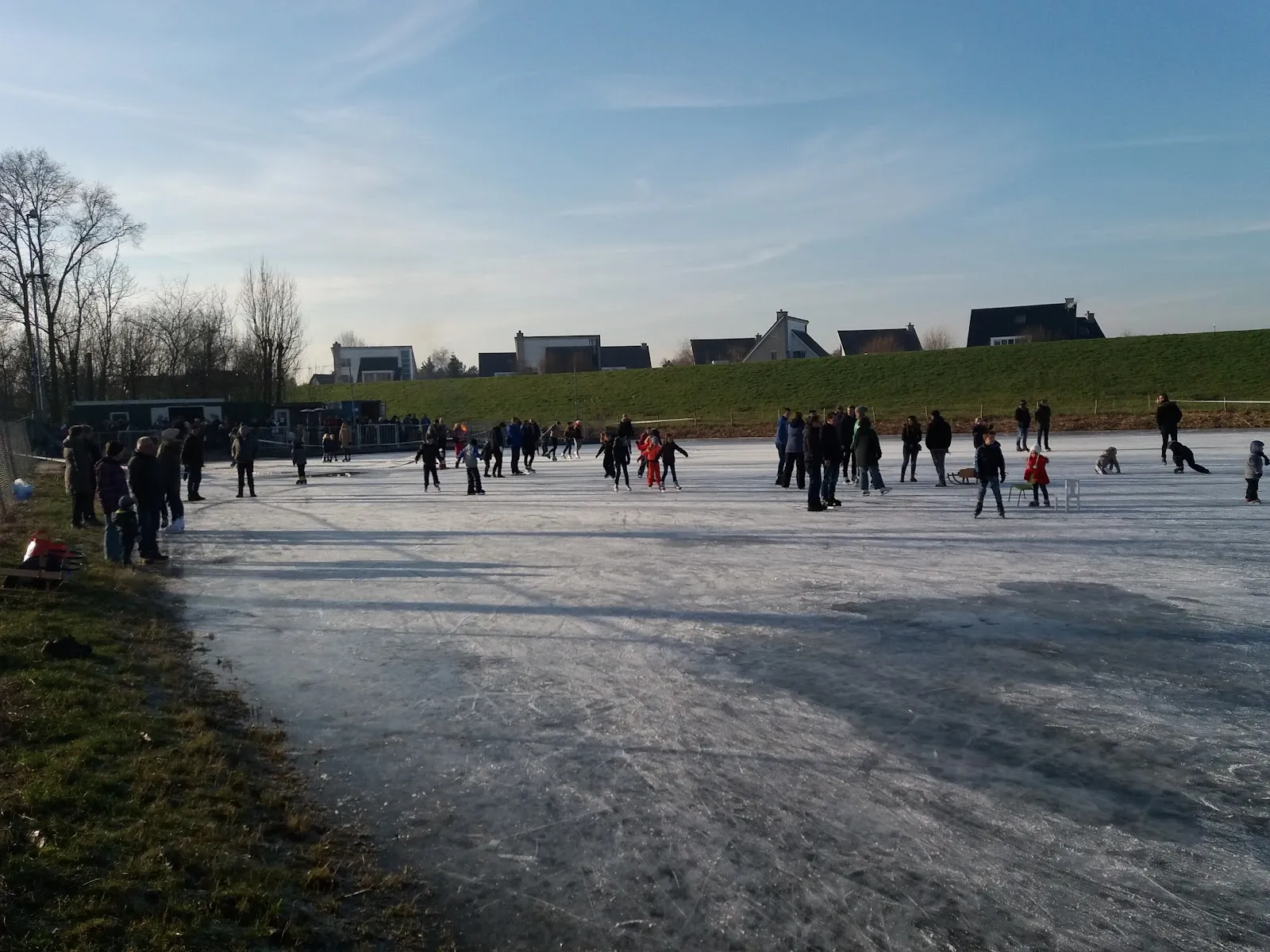 A crowd of skaters on the natural-ice rink at Terheijden beside the grassy dike, on a bright winter afternoon. — IJsclub Terheijden
