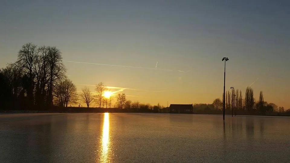 Low winter sun over the frozen rink field at Terwolde, with a floodlight pole and bare trees along the far edge. — IJsclub Terwolde