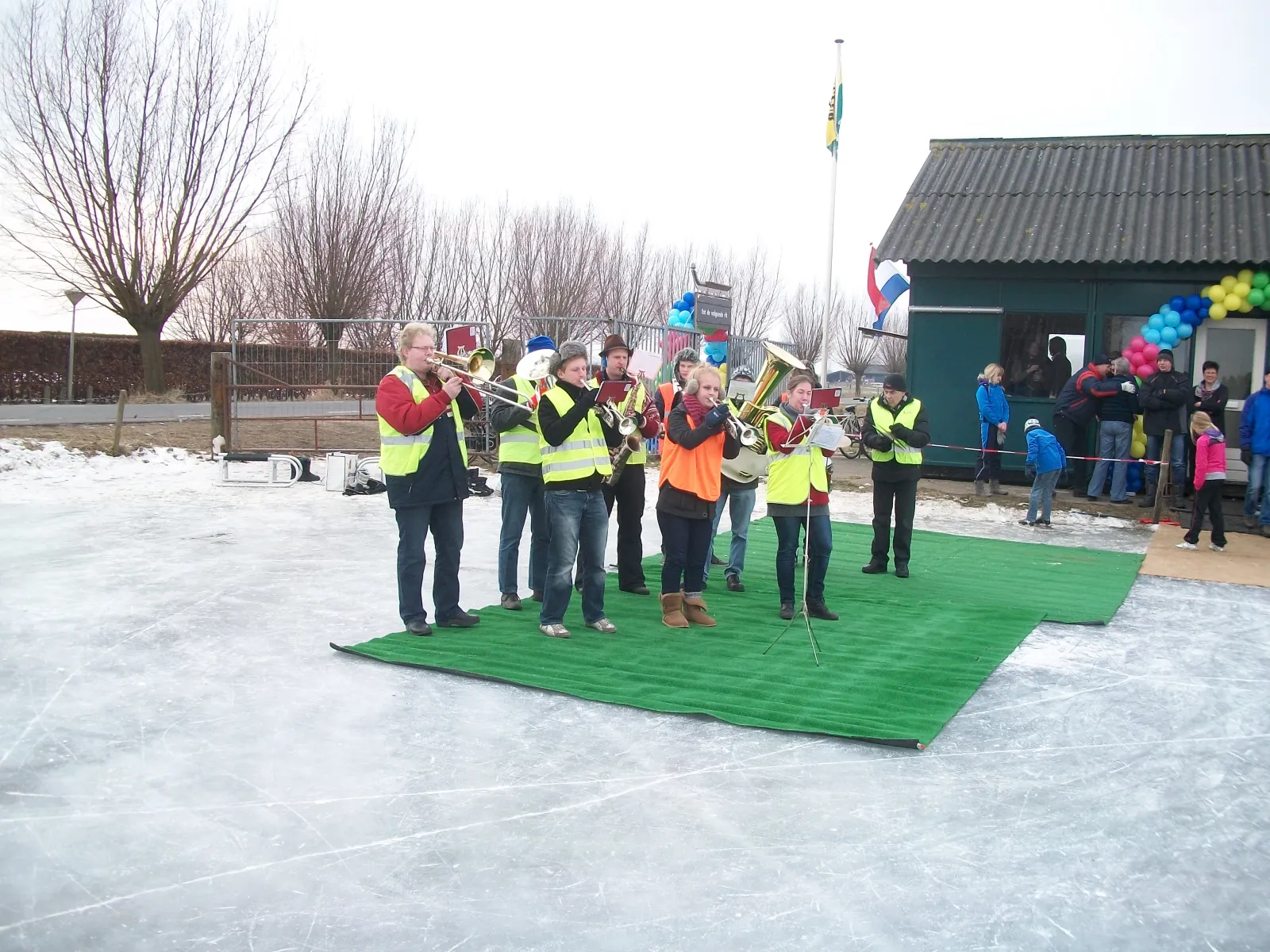 A brass band playing on a green mat on the ice beside the green clubhouse, with balloons on its roof and bare willows behind. — IJsclub Terwolde