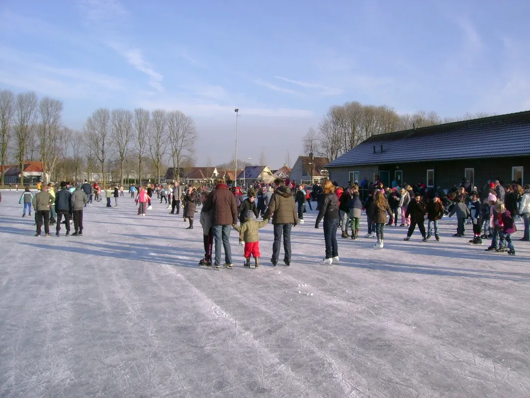 Skaters spread across the TOG rink in winter sun, with the clubhouse roofline on the right. — IJsclub TOG Kampen
