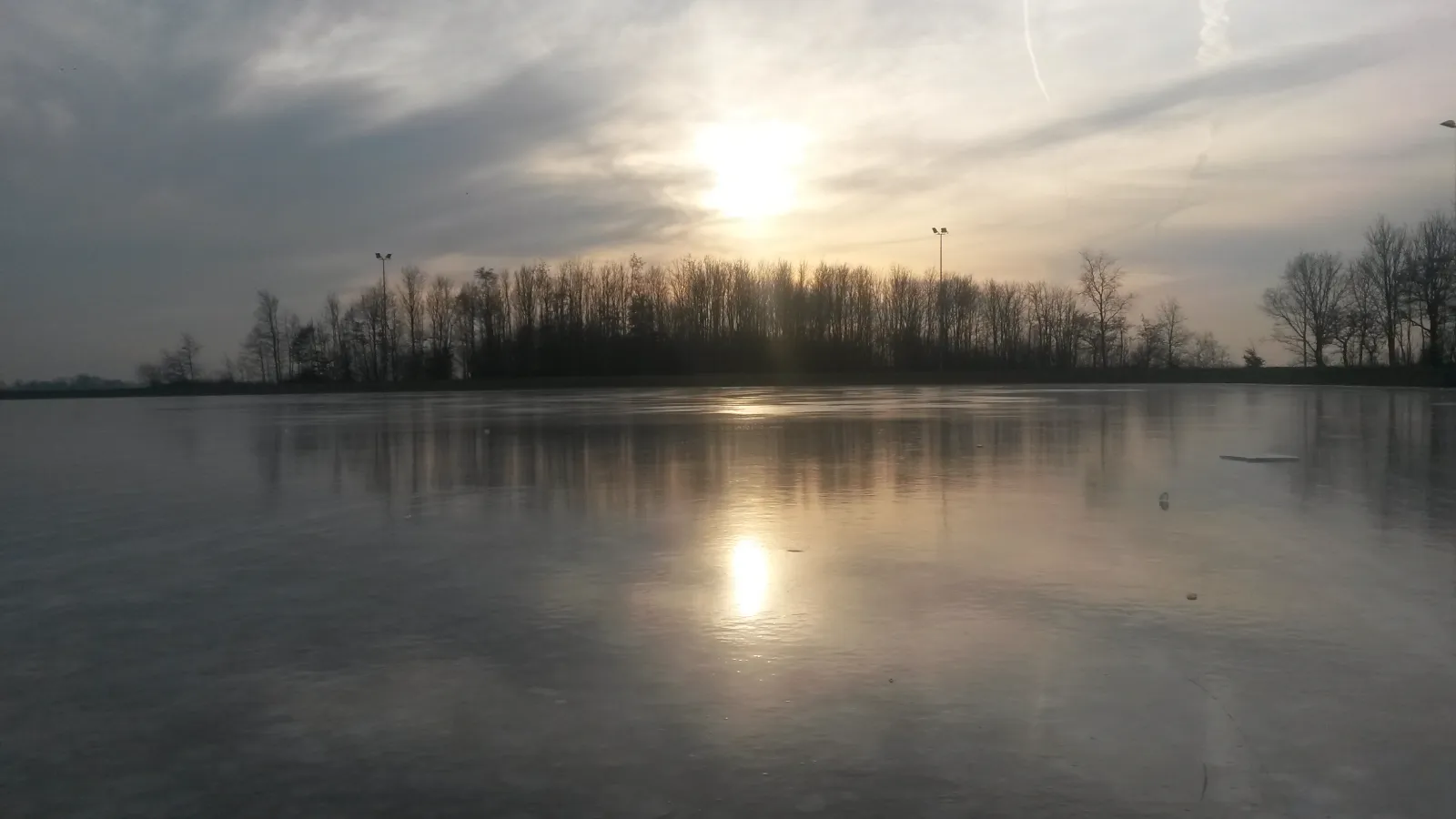 The empty natural-ice rink at Uithuizen at sunset, with the sun reflected in the smooth ice and a line of trees beyond. — IJsclub Uithuizen en omstreken