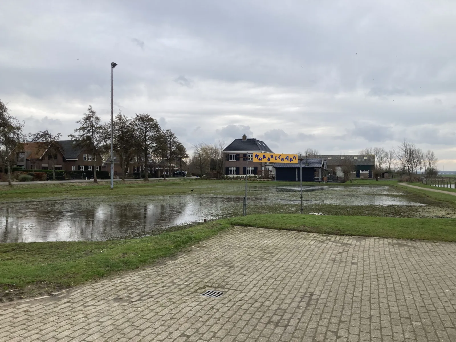 Het baangebied van Vlietland in Schipluiden met water op het vijverterrein, een bestraat plein en het clubgebouw met een geel spandoek erachter. – IJsclub Vlietland