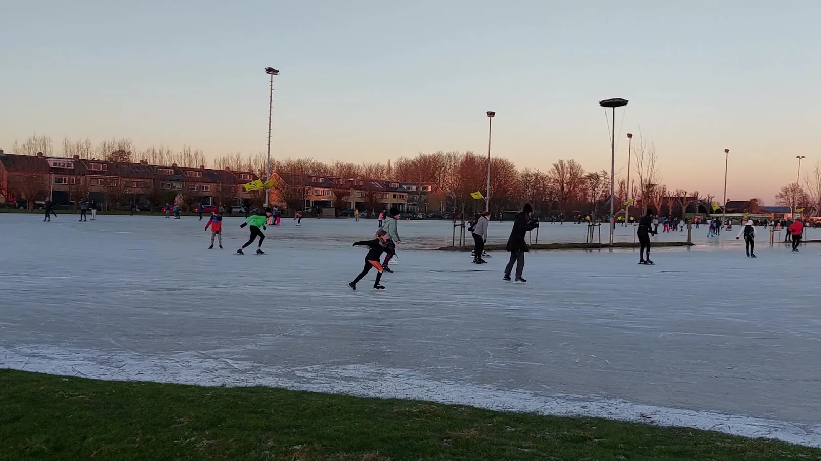 Schaatsers in de verte op de bevroren baan van Schipluiden in de schemering, met lichtmasten en de huizen van het dorp erachter. – IJsclub Vlietland