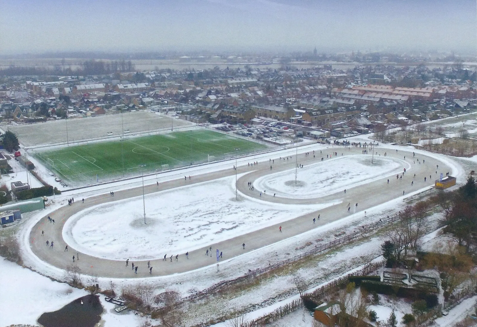 Luchtfoto van de combibaan van Ammerstol, een 200 m- en een 400 m-ovaal naast het voetbalveld, onder een dun pak sneeuw. – IJsclub Vooruitgang Ammerstol